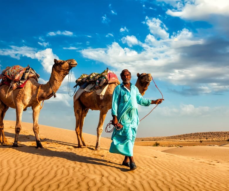 Rajasthan travel background - Indian cameleer (camel driver) with camels in dunes of Thar desert. Jaisalmer, Rajasthan, India